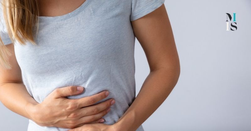 Blonde woman in gray tee shirt holds abdomen due to gallbladder removal side effects. MIIS logo at top right.